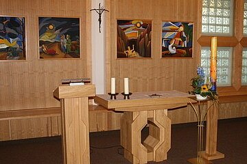 Kleine Kapelle mit hellem Holzaltar, zwei Kerzen und Lesepult. Daneben eine große Osterkerze mit Blumen. An der Wand vier farbige religiöse Bilder und ein Kruzifix. Schlichte, ruhige Sakralraumgestaltung.