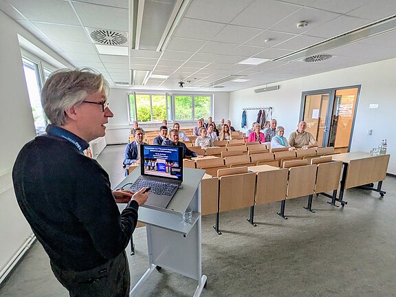 Dozent steht am Pult mit Laptop und präsentiert Folien. Vor ihm sitzt eine kleine Gruppe Teilnehmender in Reihen eines Hörsaals und hört zu. Schwerpunkt: Vortragender am Rednerpult und aufmerksam zugewandtes Publikum.
