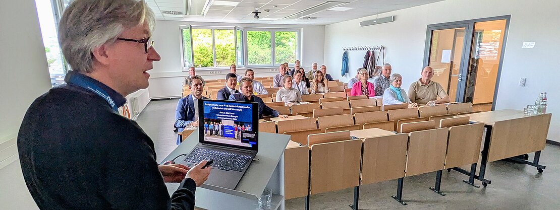 Dozent steht am Pult mit Laptop und präsentiert Folien. Vor ihm sitzt eine kleine Gruppe Teilnehmender in Reihen eines Hörsaals und hört zu. Schwerpunkt: Vortragender am Rednerpult und aufmerksam zugewandtes Publikum.