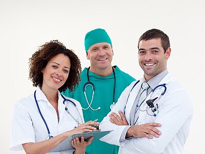 Drei medizinische Fachkräfte stehen lächelnd vor einem hellen Hintergrund. Eine Frau in weißem Kittel hält einen Notizblock, während zwei Männer, einer im grünen Operationsanzug und einer im weißen Kittel, sie unterstützen. Alle tragen Stethoskope.