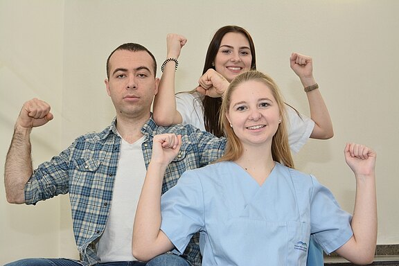 Drei junge Erwachsene posieren mit erhobenen Armen in einer kraftvollen Geste. Der Mann links trägt ein kariertes Hemd, während die Frauen in hellen T-Shirts und einer blauen Uniform gekleidet sind. Sie strahlen Selbstbewusstsein und Teamgeist aus. Der Hintergrund ist neutral gehalten.