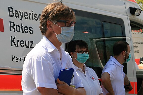Drei Mitarbeiter des Bayerischen Roten Kreuzes stehen vor einem Rettungswagen. Sie tragen Mund-Nasen-Schutz und haben eine aufmerksame Haltung. Zwei von ihnen halten Unterlagen, während der dritte nachdenklich schaut. Im Hintergrund sind Teile des Rettungswagens sichtbar.