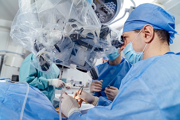 Chirurgenteam im OP führt eine präzise Mikrochirurgie durch. Der leitende Chirurg blickt durch ein steril abgedecktes Operationsmikroskop, während Hände Instrumente am Patienten führen. Schwerpunkt: moderne, hochpräzise Operation mit Teamarbeit und steriler Umgebung.