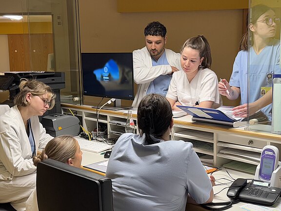 Eine Gruppe von vier Personen in Krankenhauskleidung steht um einen Schreibtisch. Zwei Personen arbeiten an einem Computer, während eine andere Informationen auf einem Tablet überprüft. Auf dem Tisch befinden sich Dokumente und ein Telefon. Ein Gefühl von Teamarbeit und Konzentration ist spürbar.
