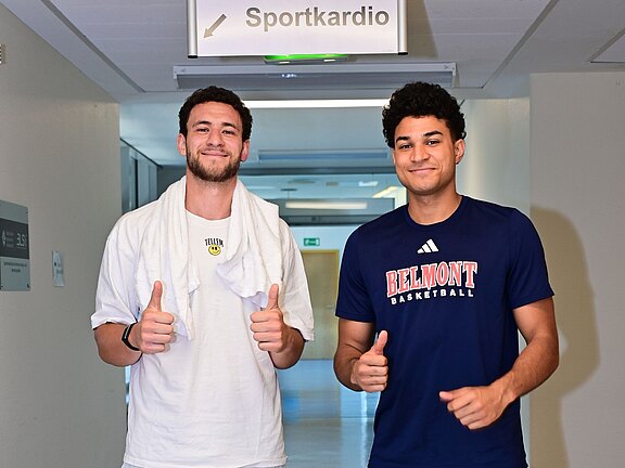 Zwei junge Männer stehen in einem hellen Flur, lächeln und zeigen beide den Daumen hoch. Links Person in Weiß mit Handtuch um den Nacken; rechts Person im dunkelblauen T-Shirt mit Aufdruck Belmont Basketball. Die Atmosphäre ist freundlich und locker.