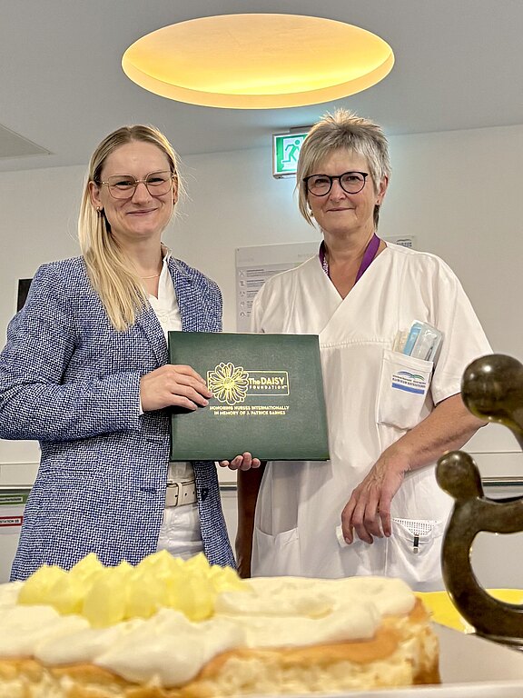 Eine Person im Anzug hält eine Mappe mit DAISY-Award-Logo neben einer lächelnden Pflegekraft im Kittel; beide blicken in die Kamera.