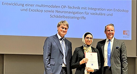 Drei Personen in formeller Kleidung stehen auf einer Bühne; die Person in der Mitte hält eine Urkunde. Im Hintergrund eine Präsentationsfolie mit dem Titel zur Entwicklung einer multimodalen OP-Technik mit Integration von Endoskop, Exoskop und Neuronavigation für vaskuläre sowie Schädelbasis-Eingriffe.