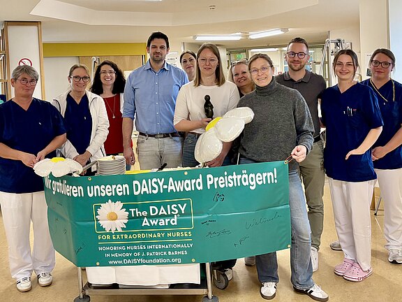 Team aus Pflegekräften und Mitarbeitenden in einem Krankenhaus posiert fröhlich mit grünem Banner The DAISY Award – For Extraordinary Nurses und Gratulation unseren DAISY-Award Preisträgern. Blumenballons in den Händen. Feierliche Szene zur Auszeichnung außergewöhnlicher Pflege.