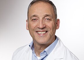 Thomas Staudacher Porträt eines lächelnden Mannes mittleren Alters in weißem Kittel und hellblauem Hemd vor neutralem Hintergrund; vermittelt Freundlichkeit und Professionalität.