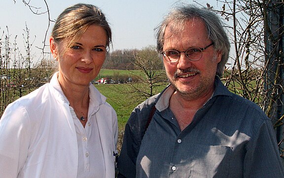 Zwei Erwachsene stehen im Freien nebeneinander. Links eine Frau in weißem Mantel, lächelnd; rechts ein Mann mit Brille und grauem Hemd, ebenfalls lächelnd. Heller Sonnenschein, unscharfe Wiese und Bäume im Hintergrund.