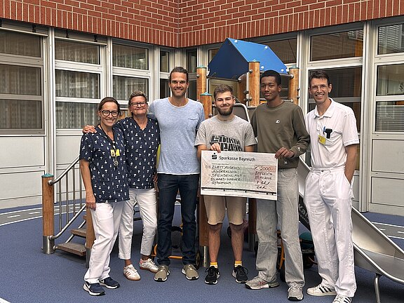 Sechs Erwachsene stehen vor einem Spielgerät und präsentieren einen übergroßen Spendenscheck der Sparkasse Bayreuth. Zwei tragen weiße Klinikbekleidung, die anderen Freizeitkleidung. Die Gruppe lächelt in die Kamera; der Scheck deutet auf eine Spendenübergabe hin.