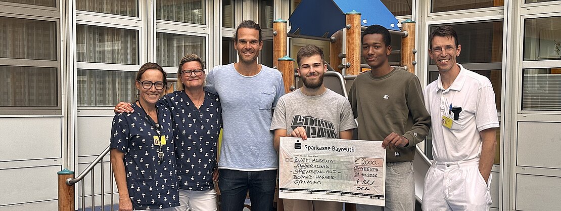 Sechs Erwachsene stehen vor einem Spielgerät und präsentieren einen übergroßen Spendenscheck der Sparkasse Bayreuth. Zwei tragen weiße Klinikbekleidung, die anderen Freizeitkleidung. Die Gruppe lächelt in die Kamera; der Scheck deutet auf eine Spendenübergabe hin.