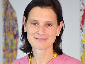 Porträt einer freundlich lächelnden medizinischen Mitarbeiterin in rosa Pflegekleidung mit Namensschild, vor neutralem Hintergrund.