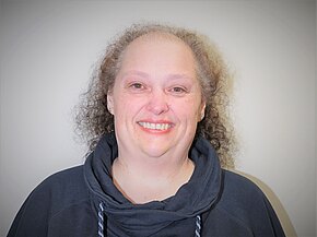 Porträt einer lächelnden Person mit schütterem, lockigem Haar. Sie trägt einen dunklen Kapuzenpullover und schaut direkt in die Kamera vor einem neutralen, hellen Hintergrund.