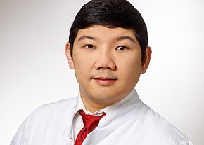 Alexander Wibisono Frontalporträt eines Mannes in weißem Kittel und roter Krawatte vor hellem Hintergrund; ruhiger, direkter Blick.