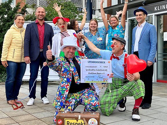 Vor einem Klinikum feiern mehrere Erwachsene; zwei Klinikclowns präsentieren einen großen Spendenscheck, zeigen Daumen hoch. Ein rotes Herz und bunte Kostüme fallen auf. Die Gruppe lächelt und jubelt; Anlass ist eine Spende zugunsten Klinikclown-Einsätze im Krankenhaus.