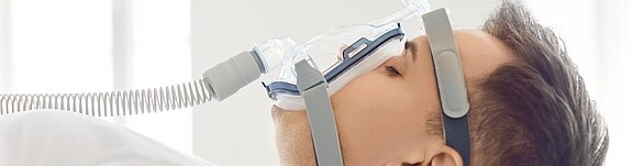 Person schläft im Bett und trägt eine CPAP-Atemmaske mit Schlauch; die Maske bedeckt Nase und Mund und ist mit Kopfbändern gesichert.