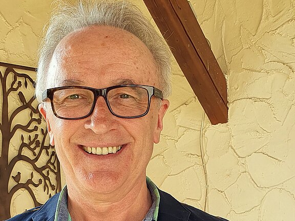 Nahaufnahme eines lächelnden Mannes mit grauem Haar und dunkler Brille. Er schaut direkt in die Kamera; sein Gesicht ist gut ausgeleuchtet. Neutraler, heller Hintergrund.