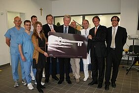 Eine Gruppe von Personen steht zusammen, die einen großen symbolischen Schlüssel präsentiert. In lässiger Kleidung sind medizinisches Personal und weitere Beteiligte zu sehen, die sich über den Schlüssel freuen. Die Szenerie vermittelt einen feierlichen Anlass, möglicherweise die Eröffnung einer neuen Einrichtung.