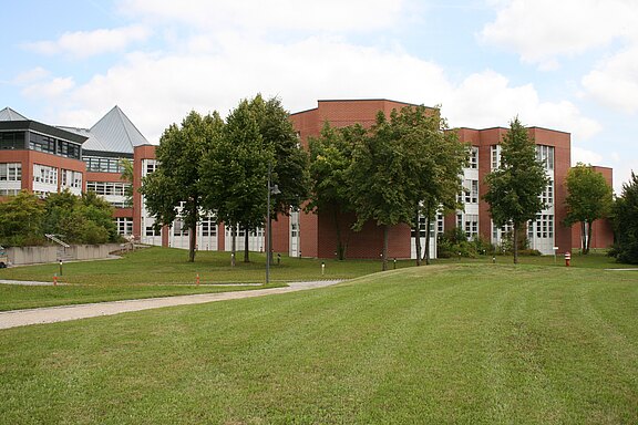 Ein modernes Gebäude aus roten Ziegeln, umgeben von Grünflächen und Bäumen. Der Himmel ist teilweise bewölkt. Auf einem Weg im Vordergrund sind Lampen aufgestellt, die den Bereich beleuchten. Das Gesamtbild vermittelt eine ruhige, einladende Atmosphäre.