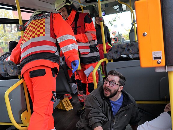Zwei Rettungssanitäter in oranger Hygieneausrüstung helfen einem verletzten Mann im Inneren eines Busses. Der Mann sitzt am Boden, wirkt überrascht und hat Schrammen im Gesicht. Eine weitere Person liegt am Boden neben ihm. Die Szene vermittelt eine Notfallsituation.
