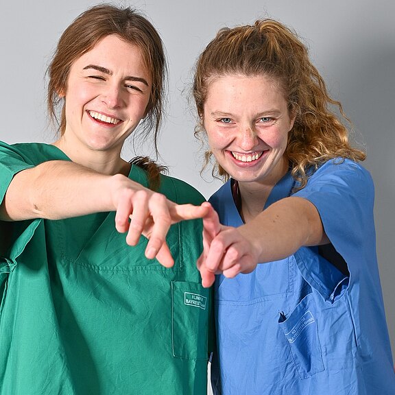 Zwei Personen in grüner und blauer medizinischer Kleidung lächeln und zeigen direkt auf die Kamera; eine fröhliche, einladende Geste.