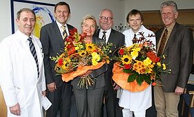 Sechs Personen stehen zusammen in einem Raum, halten große Blumensträuße mit bunten Blumen. Die Männer tragen Anzüge und der Arzt einen weißen Kittel. Die Stimmung ist festlich, da es sich um eine besondere Gelegenheit handelt.