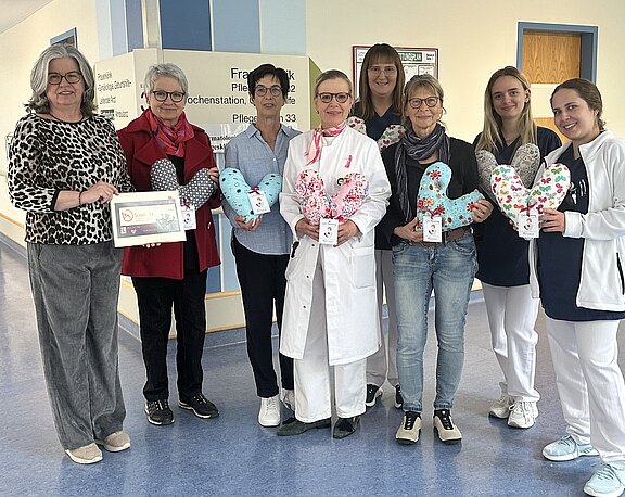 Gruppe im Krankenhausflur: Mitarbeitende und Unterstützerinnen, teils in Kittel oder Pflegekleidung, halten bunte herzförmige Kissen. Eine Person zeigt eine Urkunde. Die Szene zeigt eine Spende oder Übergabe von Trostkissen für Patientinnen auf einer Station.