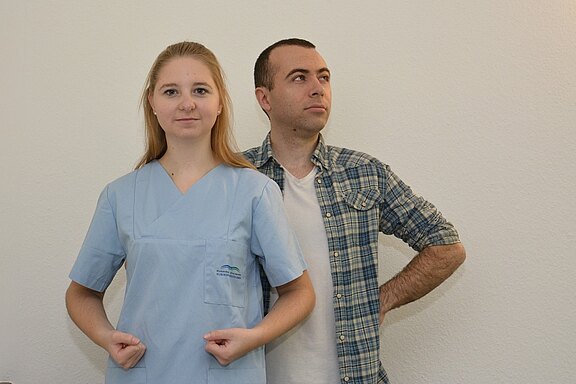 Eine junge Frau in einem hellblauen Arztkittel steht mit verschränkten Armen und einem ernsten Ausdruck, während ein Mann im Hintergrund mit zurückhaltendem Blick und lässiger Kleidung steht. Der Hintergrund ist hell und neutral.