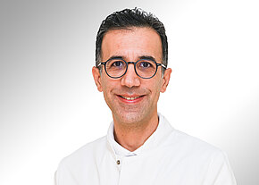 Dr. Amirfarhang Mahmoudi Porträt eines lächelnden Mannes mit dunklen, kurzen Haaren und runder Brille. Er trägt einen weißen Kittel und blickt direkt in die Kamera vor neutralem, hellem Hintergrund.