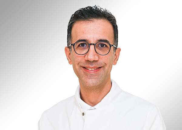 Porträt eines lächelnden Mannes mit dunklen, kurzen Haaren und runder Brille. Er trägt einen weißen Kittel und blickt direkt in die Kamera vor neutralem, hellem Hintergrund.