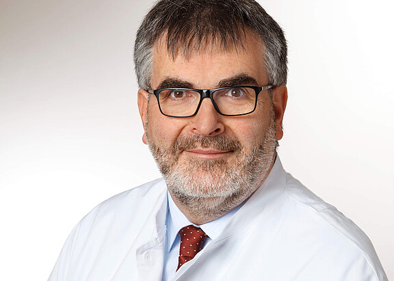 Porträt eines mittelalten Mannes mit grauem Haar und Bart, Brille, weißem Kittel und roter, gepunkteter Krawatte vor neutralem Hintergrund; er lächelt leicht und blickt in die Kamera.