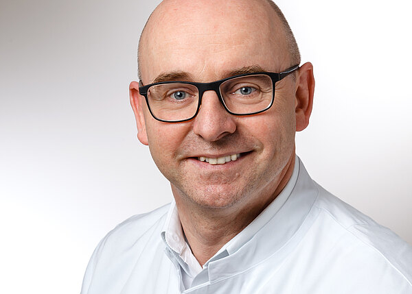 Ein lächelnder Mann mit Glatze und Brille trägt einen weißen Arztkittel. Sein Gesicht ist gut beleuchtet, und er schaut direkt in die Kamera. Der Hintergrund ist neutral und hell gehalten.