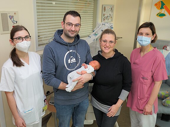 Eine Familie steht in einem Krankenhauszimmer. Der Vater hält ein Neugeborenes in seinen Armen, während die Mutter lächelt. Zwei Krankenschwestern stehen daneben, eine in weiß, die andere in pink, beide mit Mund-Nasen-Schutz. Die Umgebung vermittelt eine freundliche, fürsorgliche Atmosphäre.