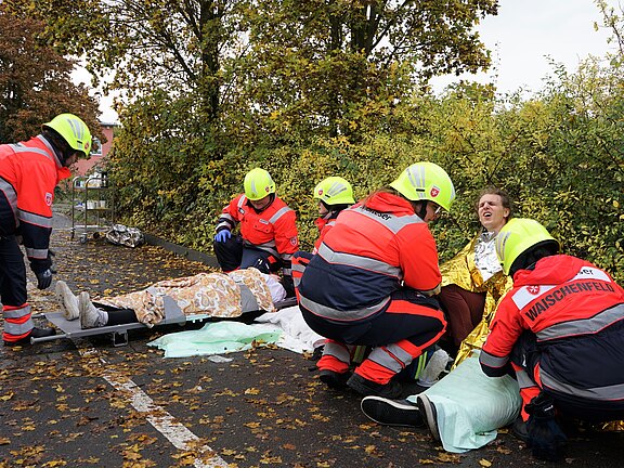 Ein Team von Rettungskräften in orangefarbener Kleidung versorgt mehrere verletzte Personen auf einer Straße, umgeben von buntem Herbstlaub. Eine Person trägt eine silberne Rettungsdecke und scheint in Schwierigkeiten zu sein. Der Fokus liegt auf der medizinischen Hilfe, die geleistet wird.
