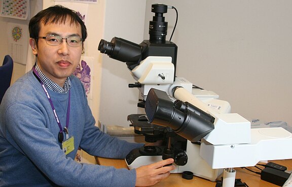 Ein Wissenschaftler in einem Labor sitzt vor einem großen Lichtmikroskop und bedient es mit der Hand. Er schaut zur Kamera. Die Szene zeigt Laborarbeit und Forschung mit mikroskopischer Analyse.