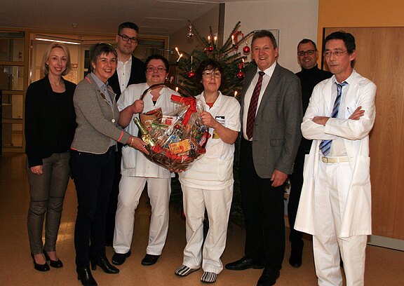 Mehrere Klinikmitarbeitende und Besucher stehen in einem Flur; zwei Pflegekräfte halten einen großen Geschenkkorb. Im Hintergrund ein geschmückter Weihnachtsbaum. Die Gruppe lächelt; es wirkt wie eine festliche Übergabe.