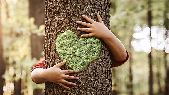 Kinderarme umschlingen einen Baumstamm. Auf der Rinde ist ein leuchtend grünes, herzförmiges Moos angebracht. Die Szene vermittelt Liebe und Schutz für die Natur.