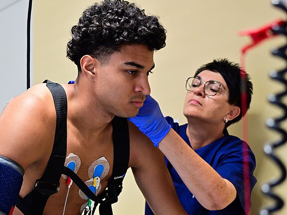 Gesundheitsfachkraft untersucht einen jungen Mann während einer sportmedizinischen Untersuchung. Er trägt Brust‑Elektroden und Gurte für Herz‑Kreislauf‑Monitoring; am Arm eine Manschette. Die Fachkraft prüft den Nackenbereich mit Handschuhen. Szene in einer Klinik, Vorbereitung oder Durchführung eines Belastungstests.