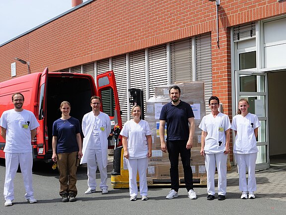 Pflegekräfte eines Krankenhauses und Helfer stehen draußen vor einem Backsteingebäude neben rotem Lieferwagen und Paletten mit Kartons; Lieferung von medizinischem Material.