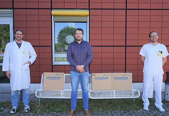 Drei Personen, zwei in weißer Klinikbekleidung, posieren vor einem Gebäude. Auf der Bank vor ihnen stehen mehrere Kartons mit der Aufschrift „OLYMPUS“ – offenbar eine Lieferung oder Übergabe von medizinischer Ausrüstung.