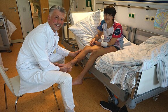 In einem Krankenhauszimmer untersucht ein Arzt einen jugendlichen Patienten, der im Bett sitzt. Der Arzt hält das Bein des Patienten, offenbar zur Kontrolle oder Physiotherapie. Der Patient trägt einen Verband am Arm und wirkt entspannt.