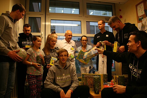 Eine Gruppe von Basketballspielern besucht eine Kinderklinik. Sie stehen mit Kindern und Klinikpersonal zusammen und überreichen Spielsachen und Bücher. Die Stimmung ist freundlich, die Kinder halten Geschenke in den Händen.
