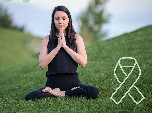 Frau in schwarzer Sportkleidung meditiert im Schneidersitz auf einer grünen Wiese; Augen geschlossen, Hände zum Gebet vor der Brust. Ruhe und Achtsamkeit.