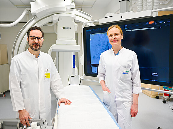 Zwei Mediziner in einem modernen Eingriffsraum mit großem Monitor und Röntgenanlage; sie stehen am Behandlungstisch und lächeln in die Kamera.