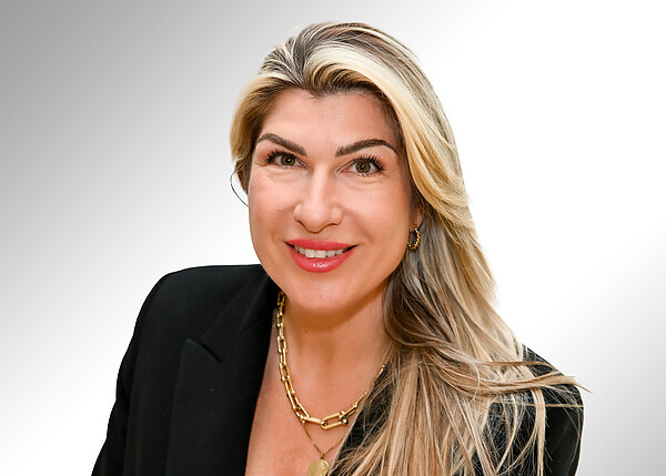 Porträt einer lächelnden Frau mit langen, blonden Haaren. Sie trägt einen schwarzen Blazer und goldene Ketten. Brustbild, direkter Blick in die Kamera vor hellem Hintergrund.