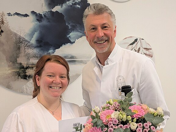 Zwei lächelnde medizinische Fachkräfte in weißen Kitteln posieren in einer Praxis. Die Person links hält ein Dokument, die Person rechts einen bunten Blumenstrauß. Die Szene deutet auf eine Gratulation oder Auszeichnung hin.