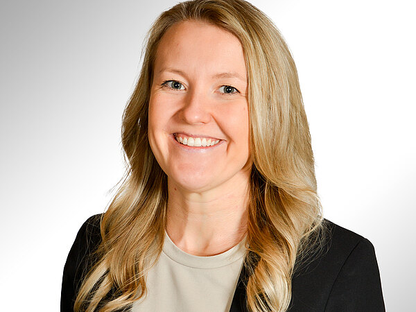 Porträtfoto einer lächelnden Frau mit langen, blonden, welligen Haaren. Sie trägt ein helles Oberteil und einen dunklen Blazer vor hellem Hintergrund.