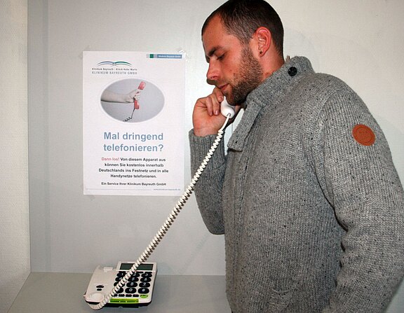 Ein Mann im grauen Pullover telefoniert mit einem weißen, schnurgebundenen Tischtelefon. Im Hintergrund hängt ein Plakat mit der Überschrift „Mal dringend telefonieren?“, das auf einen Telefonservice hinweist.