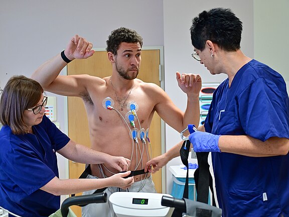 Zwei Pflegekräfte bereiten einen oberkörperfreien Mann mit Brustelektroden auf ein Belastungs-EKG vor. Sie befestigen Kabel und einen Gurt, der Mann hebt die Arme; medizinisches Untersuchungsgerät steht im Vordergrund.